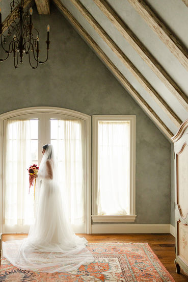 Bride in a lace gown gazing out an arched window in a timber-beamed chateau bedroom, holding an autumn bouquet
