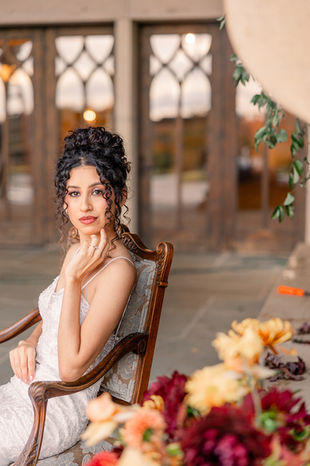Bride with a curled updo seated in a vintage wooden chair beside autumn florals