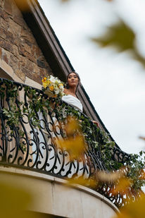 Bride on a wrought-iron balcony overlooking the grounds, framed by autumn leaves