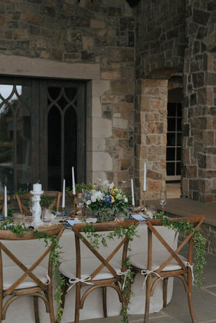 Outdoor dining table set with candles and greenery beside cross-back wooden chairs