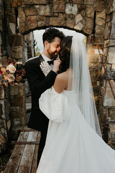 Bride and groom embracing beneath a stone archway, foreheads touching