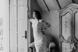 Bride reaching upward in a sunlit doorway beside an antique carved armoire